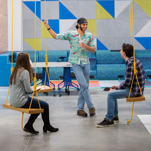 At the Hatchery, a student stands and talks with two students sitting on swings