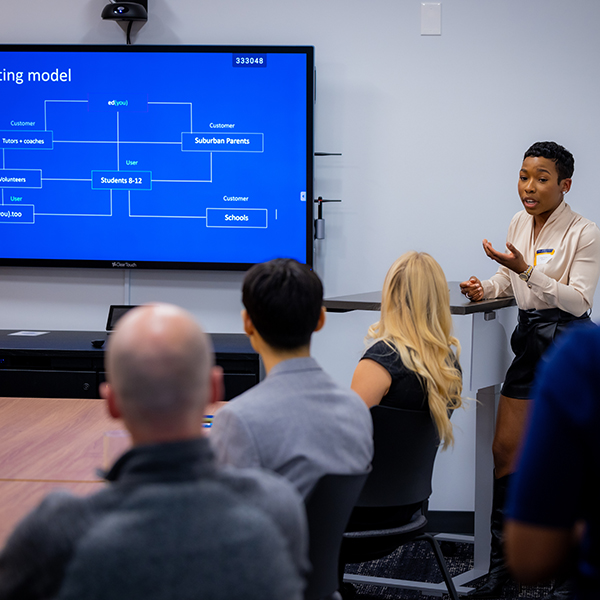 A young woman next to a large screen, giving a presentation to a group