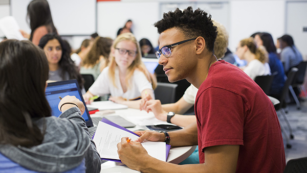 A student taking notes on a document has paused to listen to his classmate while a class full of students is visible in the background