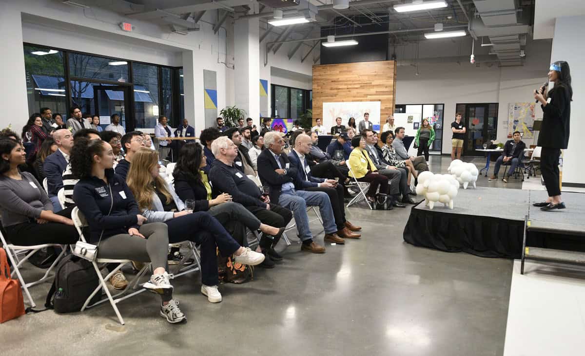 a full audience listening to a woman speaking on stage behind two toy sheep