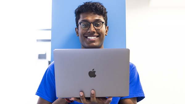 A young man smiles from behind his laptop screen